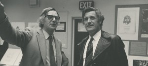 A black and white image of two men. The person on the left is former Festival Director Michael Dawson. He wears a grey suit with a tie, glasses and is pointing to his left. The person on the right is poet Ted Higher. He wears a black jacket with a tie and is looking up to his left. They stand in the King’s Hall, with a door and posters in the background.