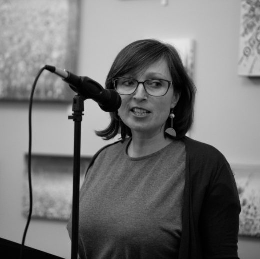 An image of a woman with short, dark hair, stood in front of a microphone.