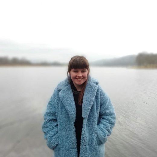 An image of a woman wearing a blue coat stood in front of water.