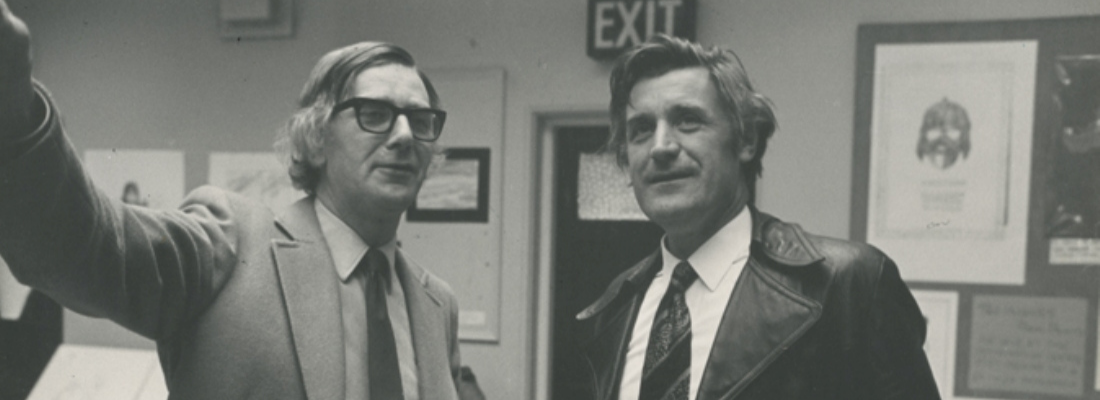 A black and white image of two men. The person on the left is former Festival Director Michael Dawson. He wears a grey suit with a tie, glasses and is pointing to his left. The person on the right is poet Ted Hughes. He wears a black jacket with a tie and is looking up to his left. They stand in the King’s Hall, with a door and posters in the background.