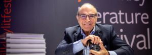 Image of David Suchet smiling, holding a camera, with a pile of books